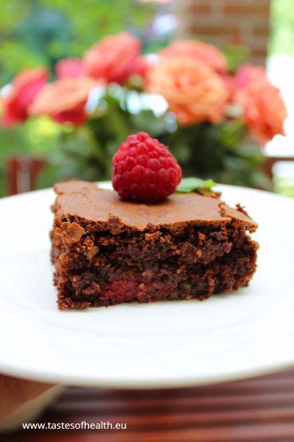 An image of Gluten Free Brownies on a white plate with a raspberry on top. There are peach-coloured roses in the background.