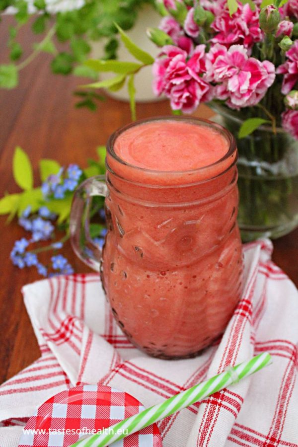 An image of Strawberry and Pineapple Smoothie in a glass on a white and red teatowel. There are blue and pink flowers in the background.