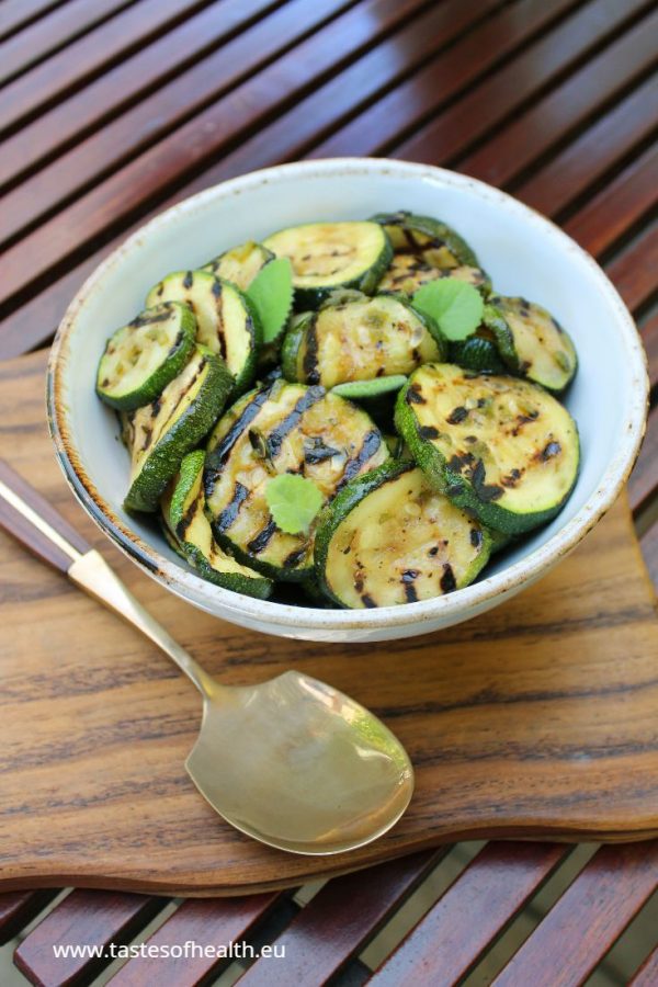 An image of Courgette Salad in a white bowl standing on a wooden board. The board is on a table.