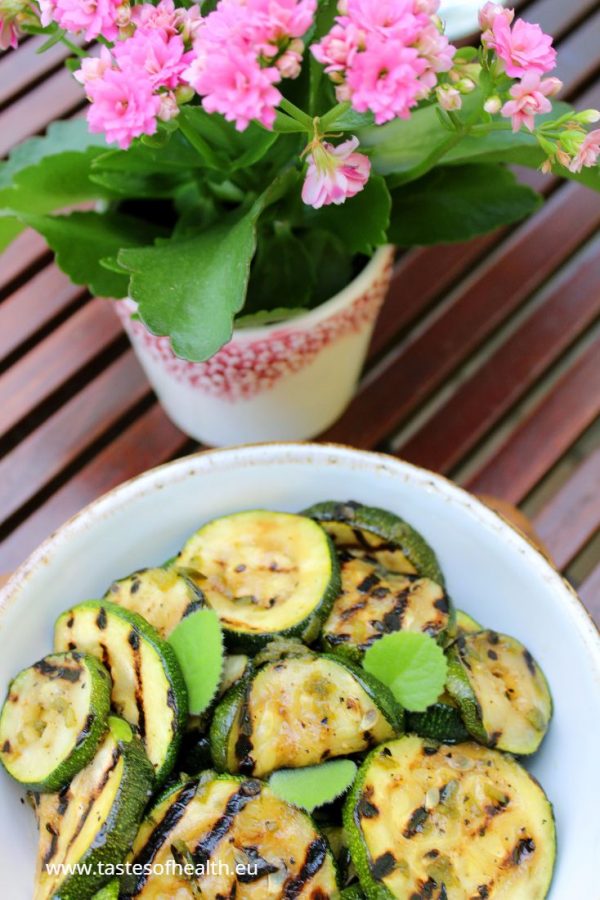 An image of Courgette Salad in a white bowl standing on a wooden board. The board is on a table and there is a pot with pink flowers next to it.