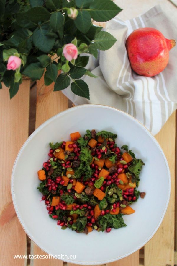 Roasted Kale Salad with Spiced Roasted Pumpkin in a white bowl. There is a blooming pink rose, a pomegranate and a beige cloth next to the bowl.