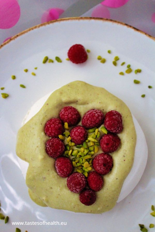 An image for the post: Vegan Meringue Recipe. The aquafaba meringue has pistachio cream and raspberries arranged in a shape of a heart on top. The meringue is on white plate standing on a piece of fabric with pink dots.